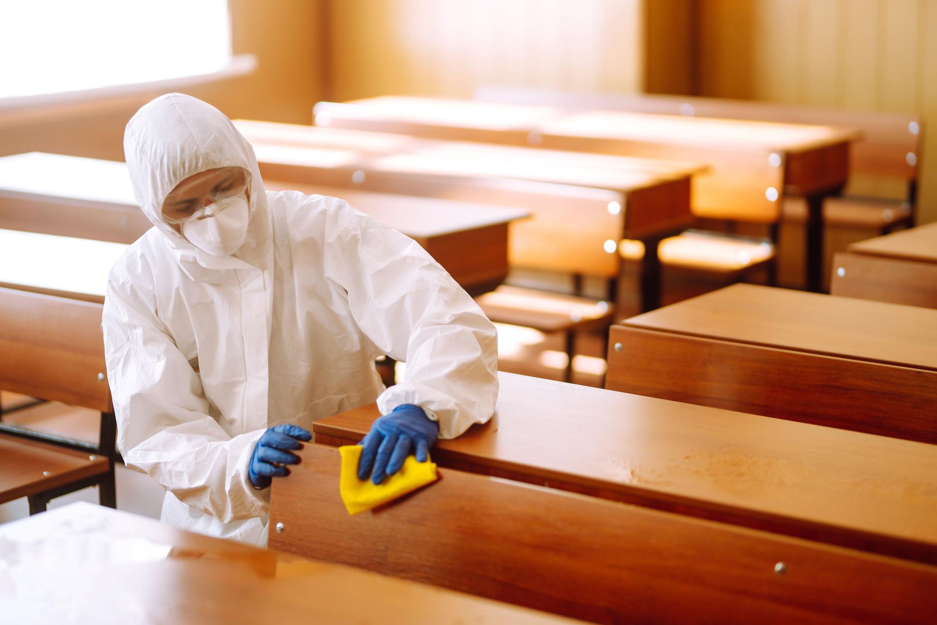 Man in a protective suit washes school desk.